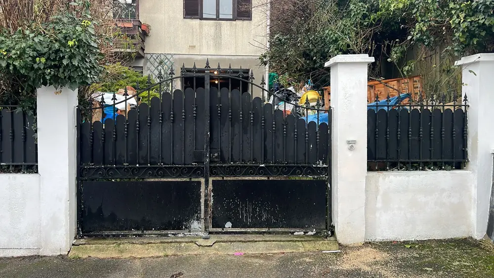 Portail battant en acier noir entre deux piliers blancs, devant un pavillon